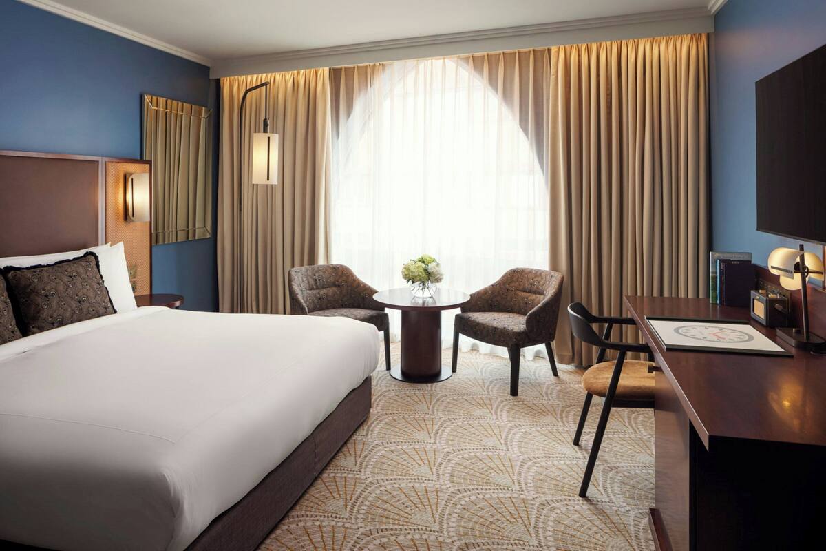 A hotel room with a large bed, a round table with two chairs, a desk with a chair, a TV, and a large window with curtains.