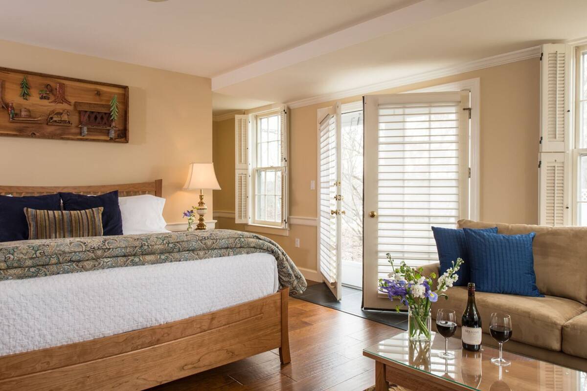 Um quarto aconchegante com uma cama de madeira, uma mesa de centro de vidro com uma garrafa e um copo, e um sofá com almofadas.