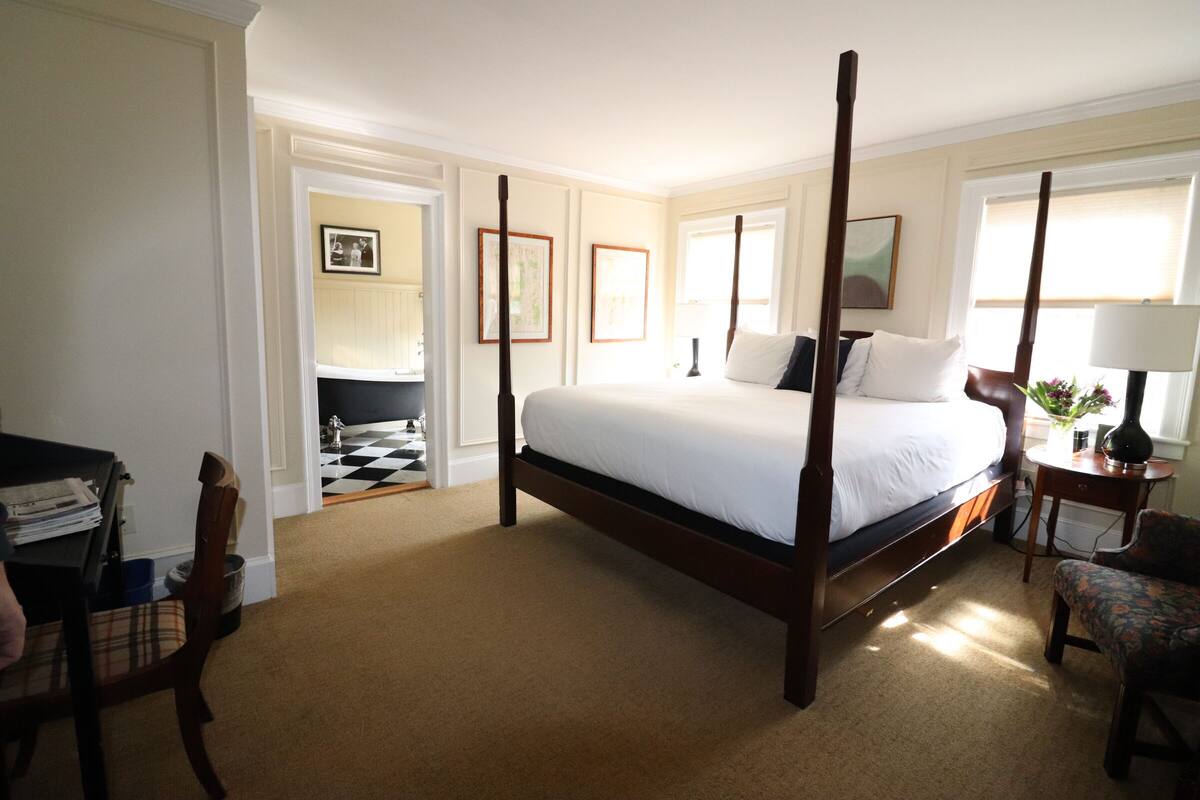 Uma cama de dossel em quarto de hotel com piso xadrez, uma escrivaninha com papéis e uma cadeira.