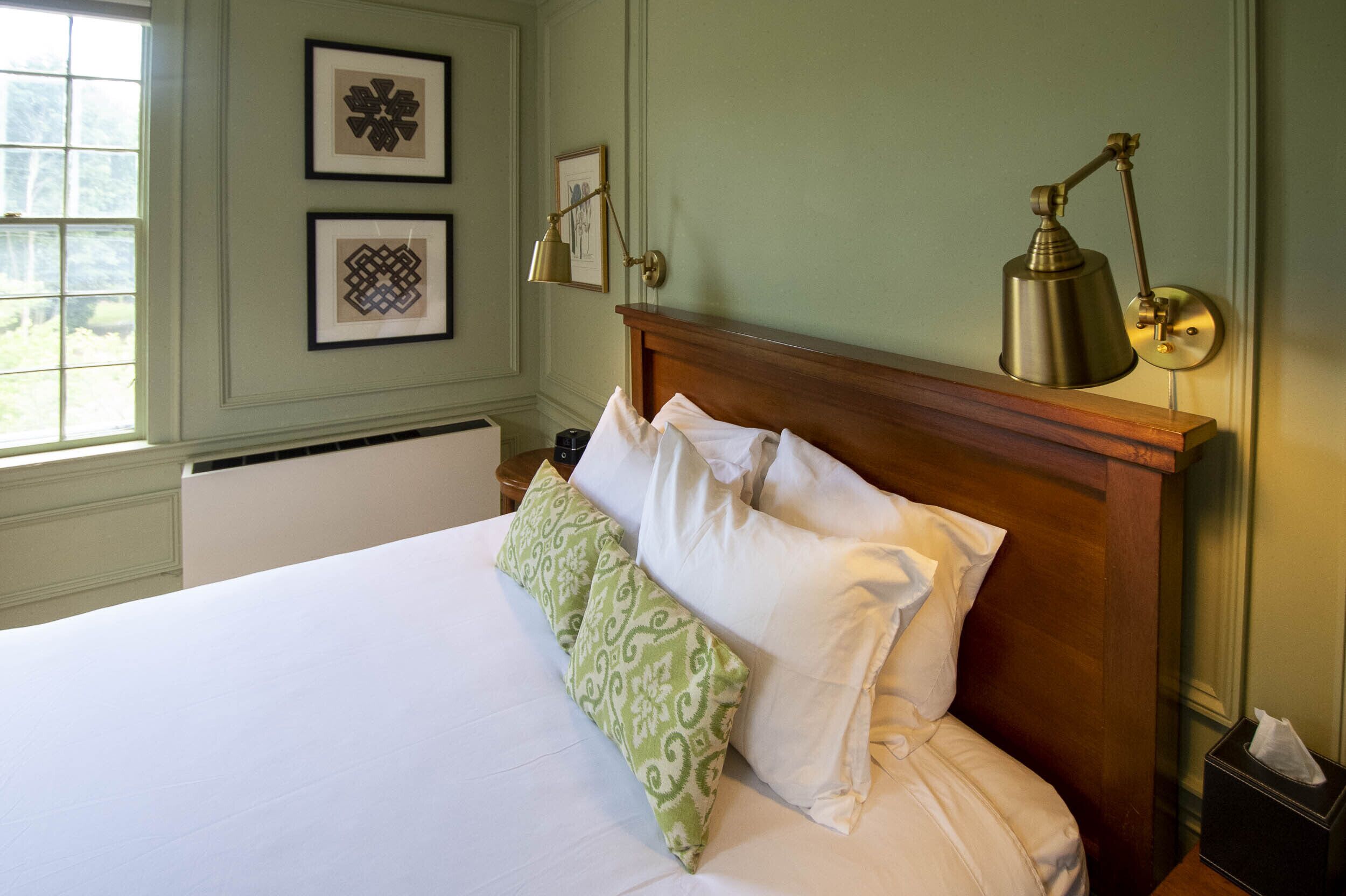 A neatly made bed with white and green pillows, a wooden headboard, and a brass desk lamp on a bedside table.