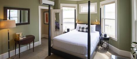 A four-poster bed with white linens, a bedside table with a lamp, and a window with a view of the outdoors.