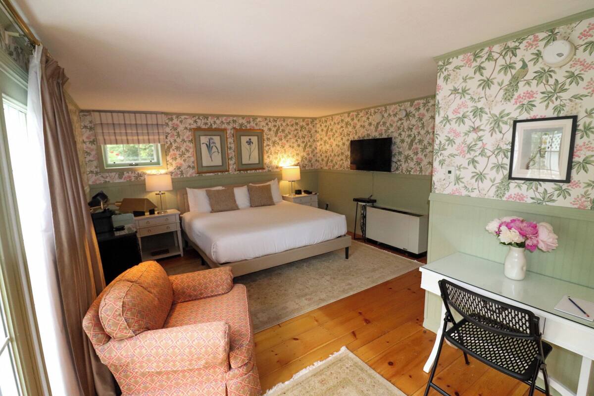 Quarto com cama, mesas de cabeceira, escrivaninha com cadeira, televisão e papel de parede floral.