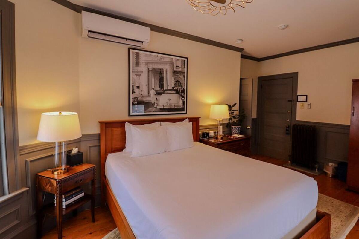 Quarto de hotel com uma cama grande, duas mesas de cabeceira com luminárias, um aparelho de ar condicionado, um quadro pendurado na parede, uma porta e um guarda-roupa.