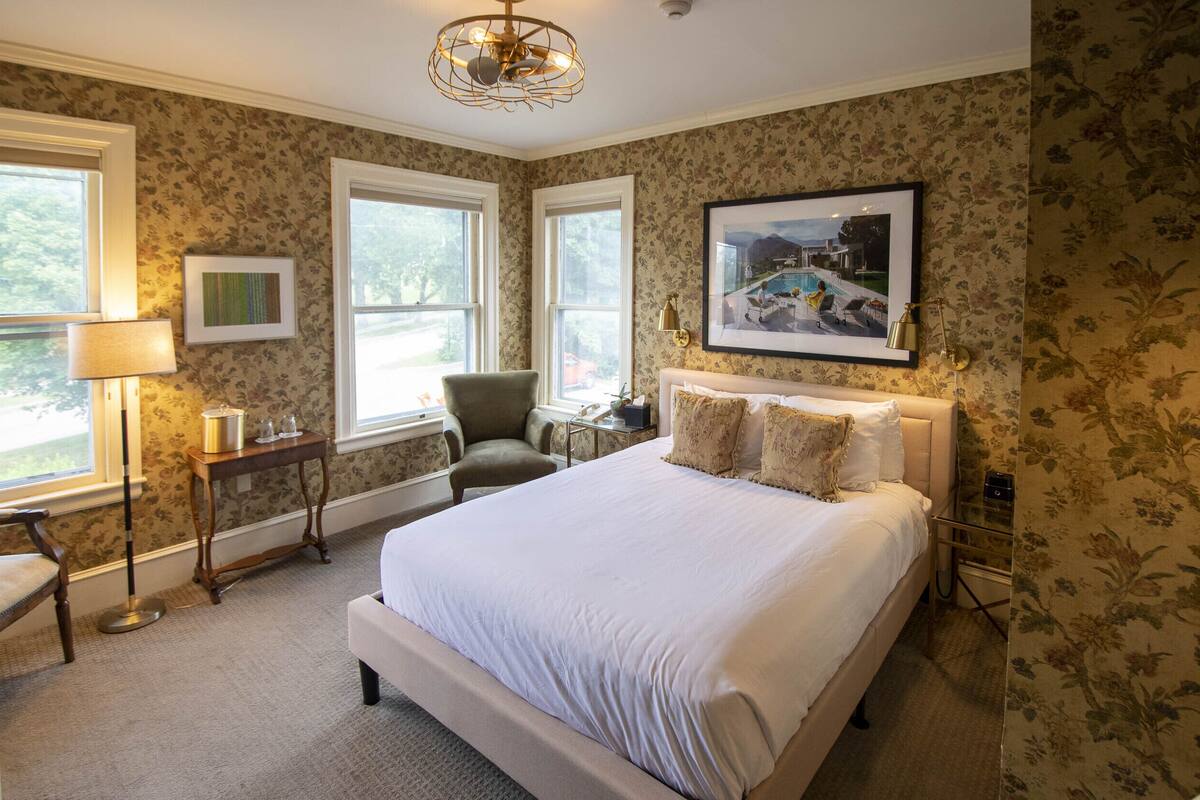 Quarto com uma cama grande, duas janelas, uma cadeira, uma mesa e um quadro na parede.