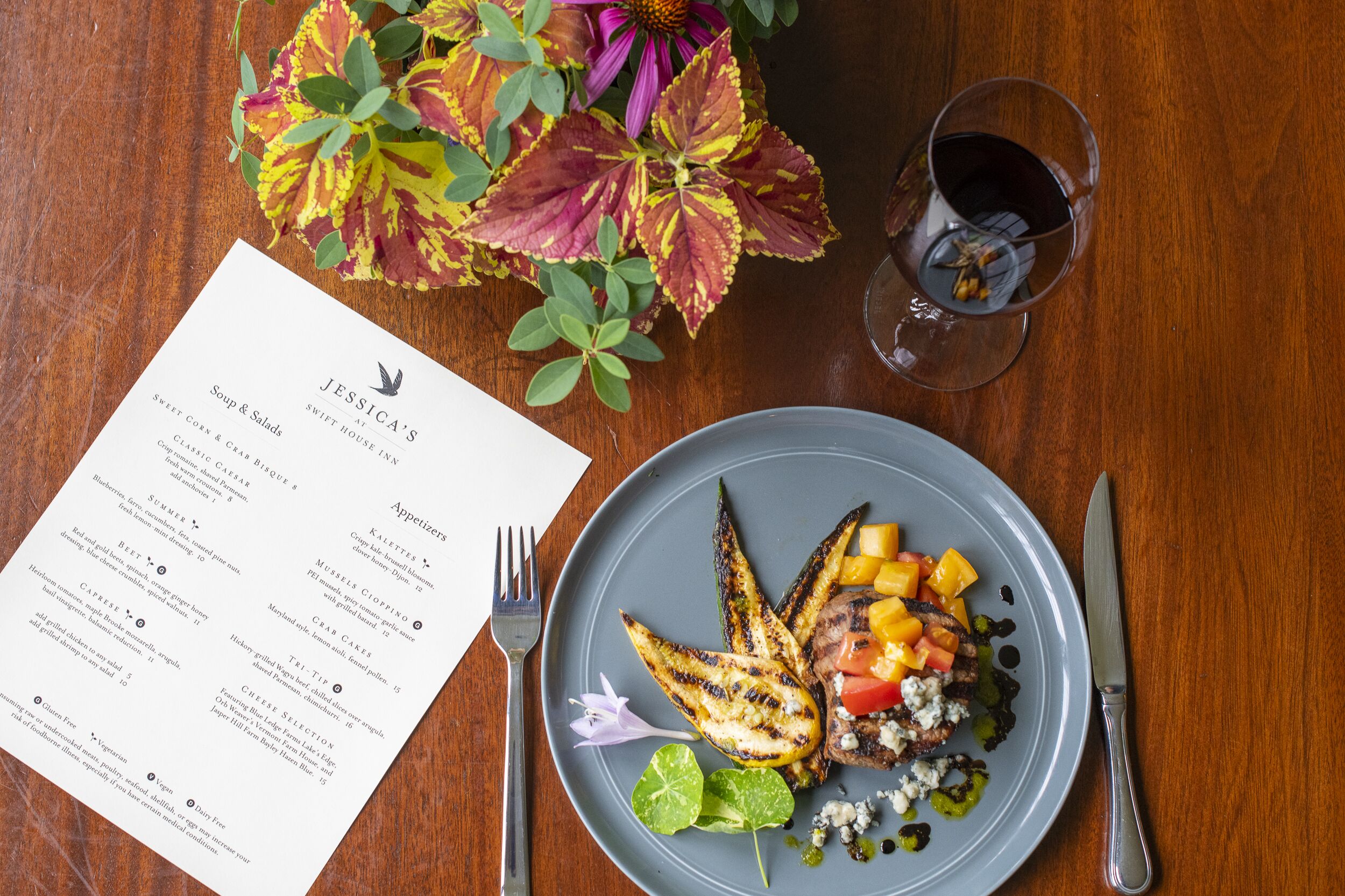 Un menu con diverse portate, un piatto con verdure e carne alla griglia, un bicchiere di vino rosso e una forchetta e un coltello su un tavolo di legno.