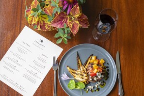 A menu with a variety of dishes, a plate with grilled vegetables and meat, a glass of red wine, and a fork and knife on a wooden table.