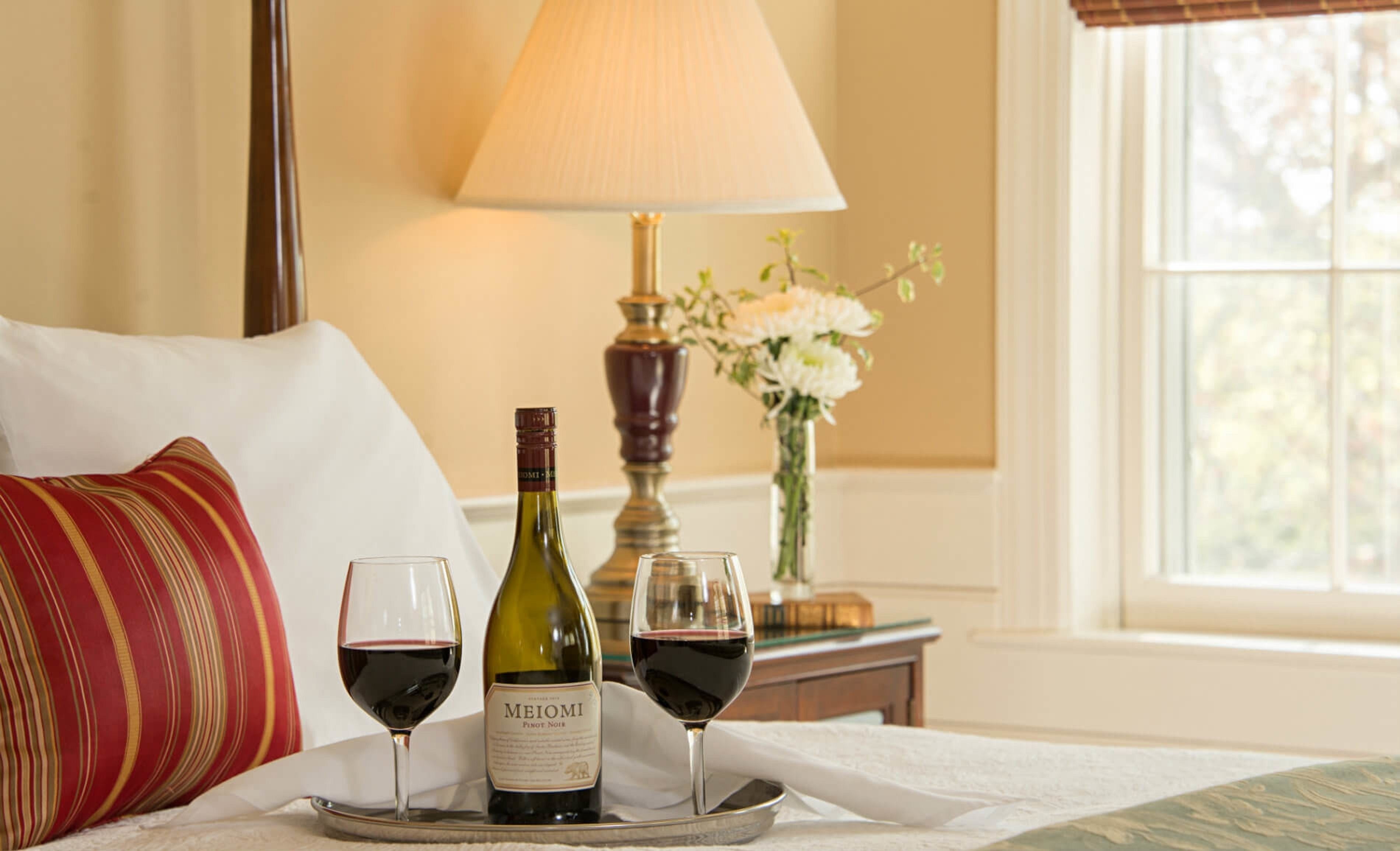 A bed with a pillow, a tray with a wine bottle and two glasses of red wine, a lamp, and a vase with flowers.