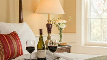 A bed with a pillow, a tray with a wine bottle and two glasses of red wine, a lamp, and a vase with flowers.