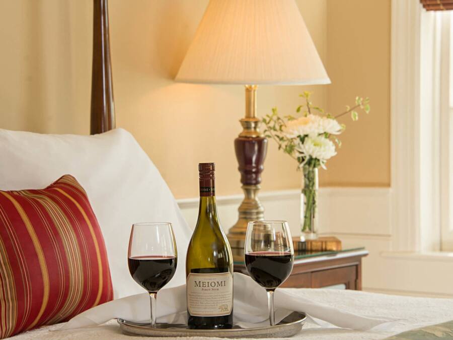 A bed with a pillow, a tray with a wine bottle and two glasses of red wine, a lamp, and a vase with flowers.