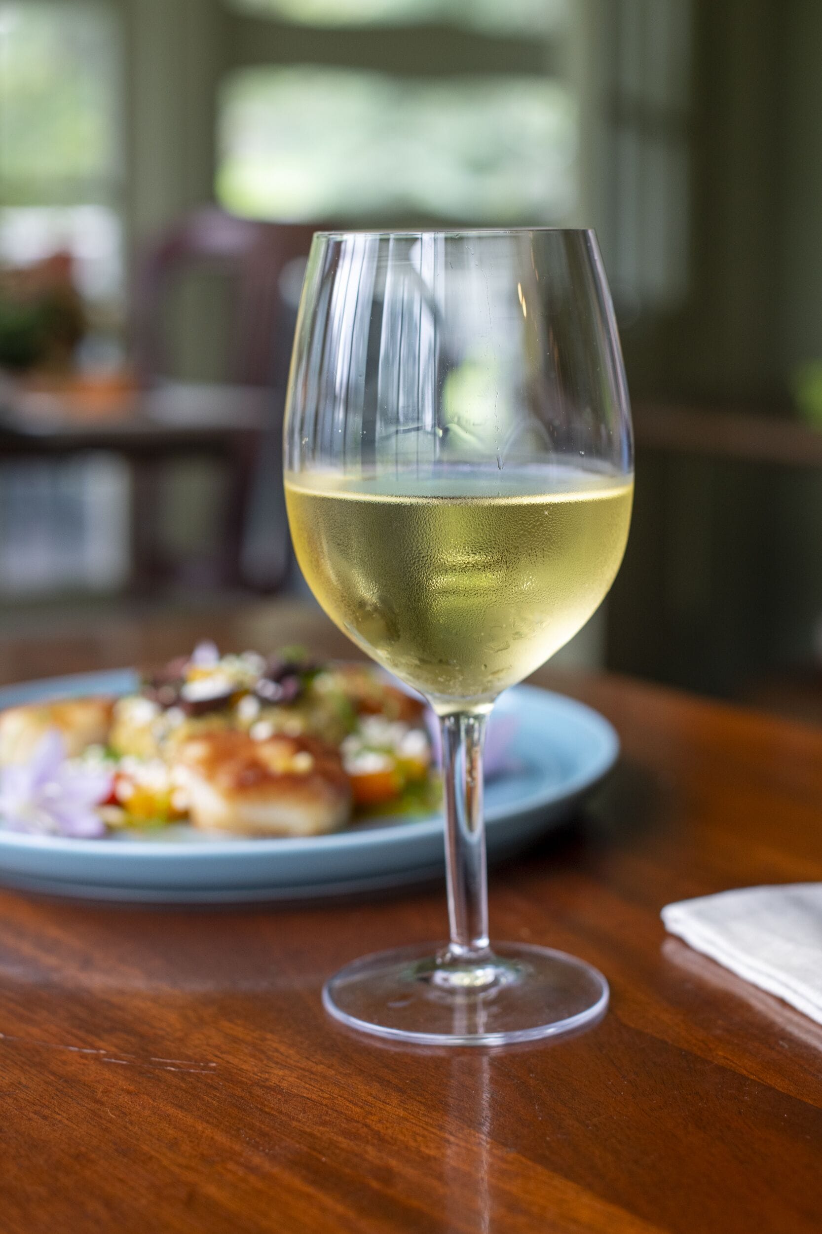 A glass of white wine on a wooden table with a plate of food in the background.