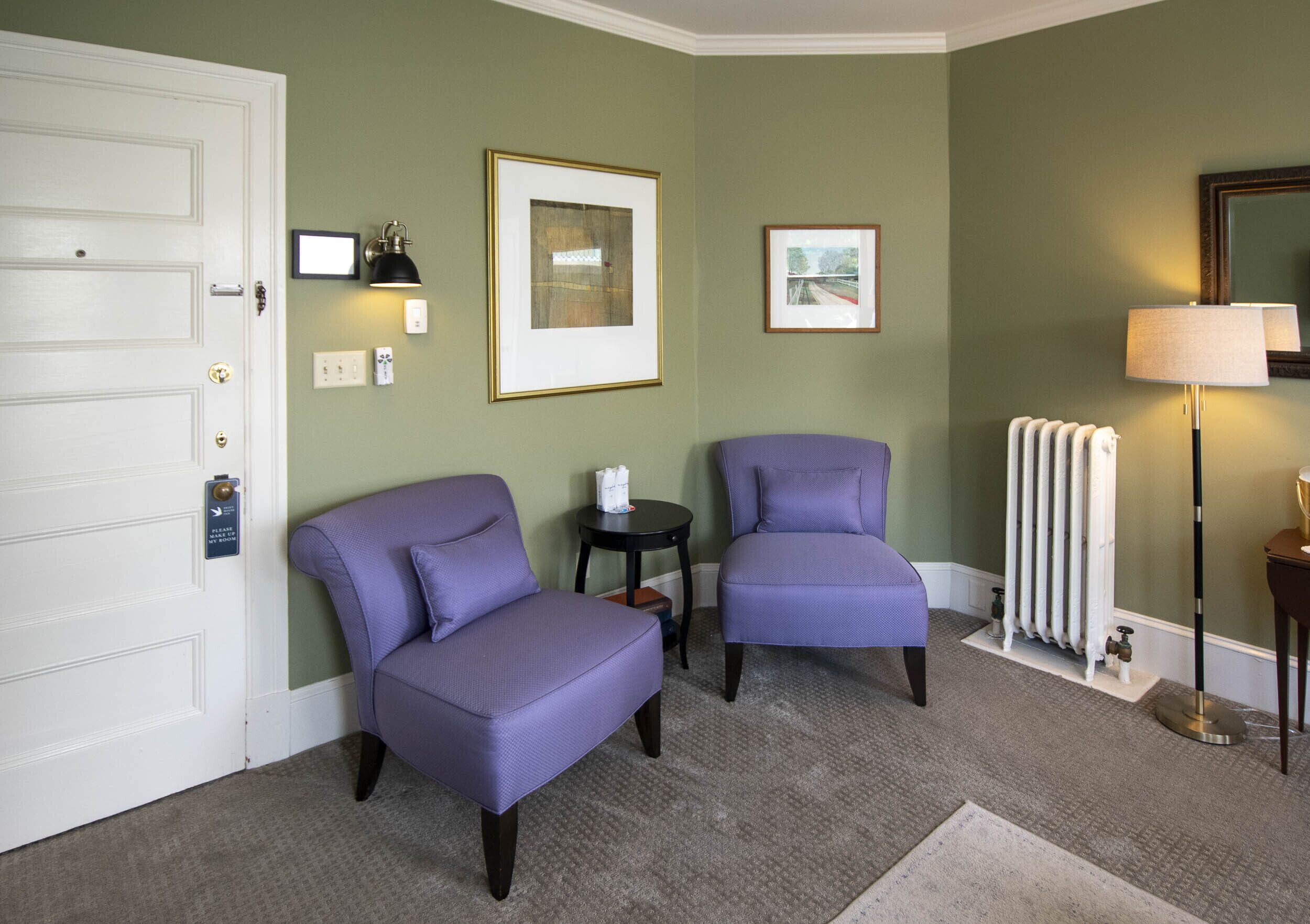 A room with two purple armchairs, a white door, a radiator, and framed pictures on the wall.
