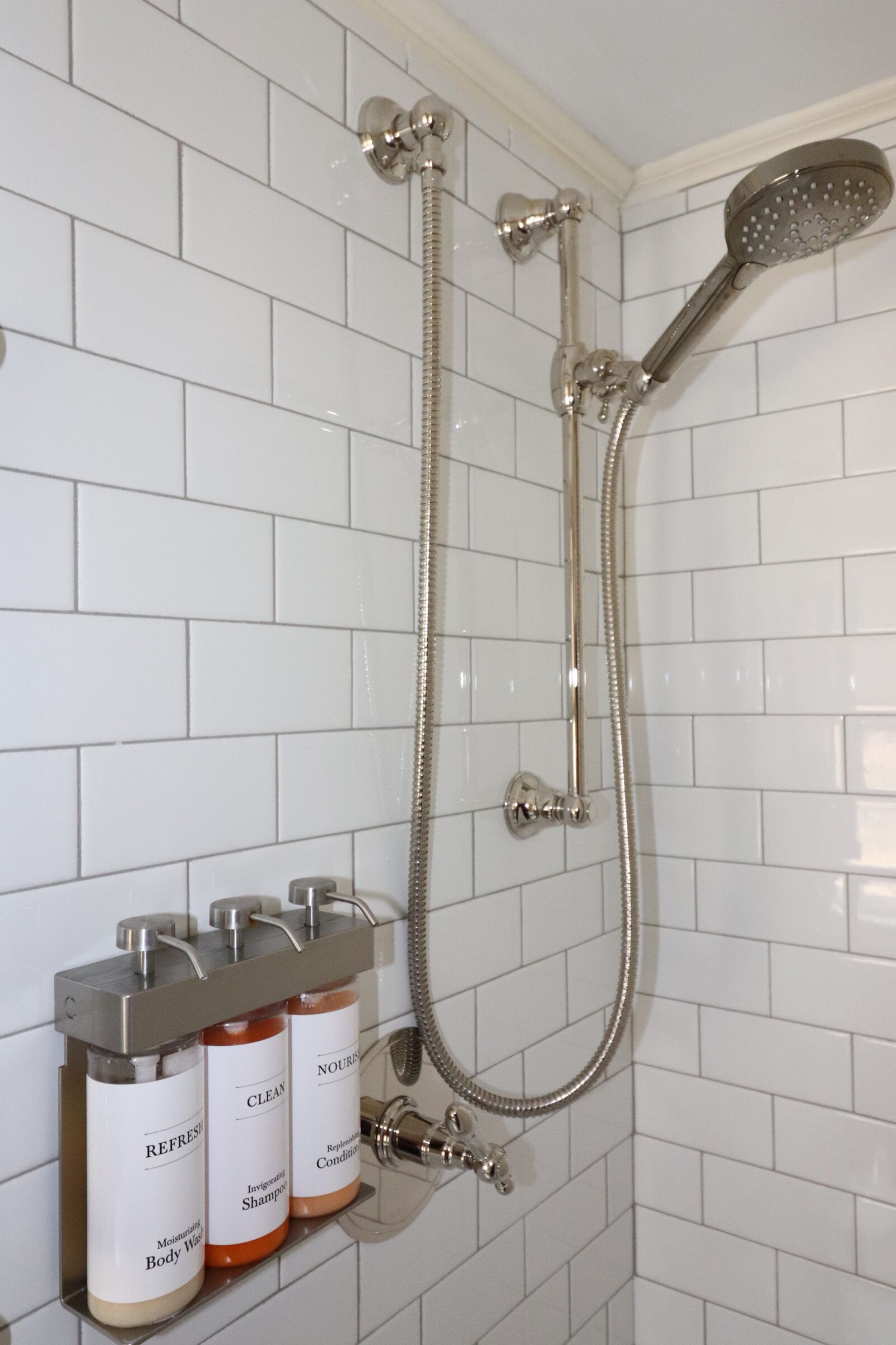 Un baño con paredes de azulejos blancos, una ducha con cabezal de mano y tres botellas de gel de baño y champú sobre una estantería.
