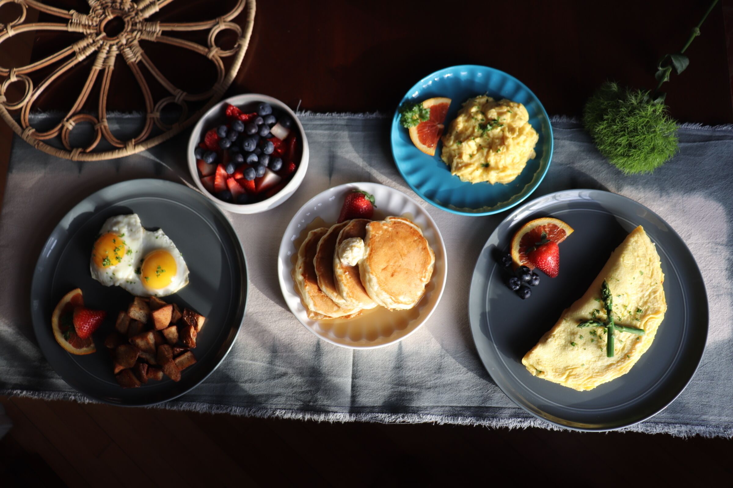 Una mesa de desayuno con platos de huevos, panqueques y una tortilla, acompañada de frutas y tostadas.