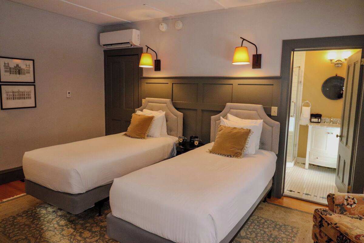 Quarto de hotel com duas camas de solteiro, uma mesa pequena e uma cadeira. Há uma porta que dá para um banheiro com espelho e pia.