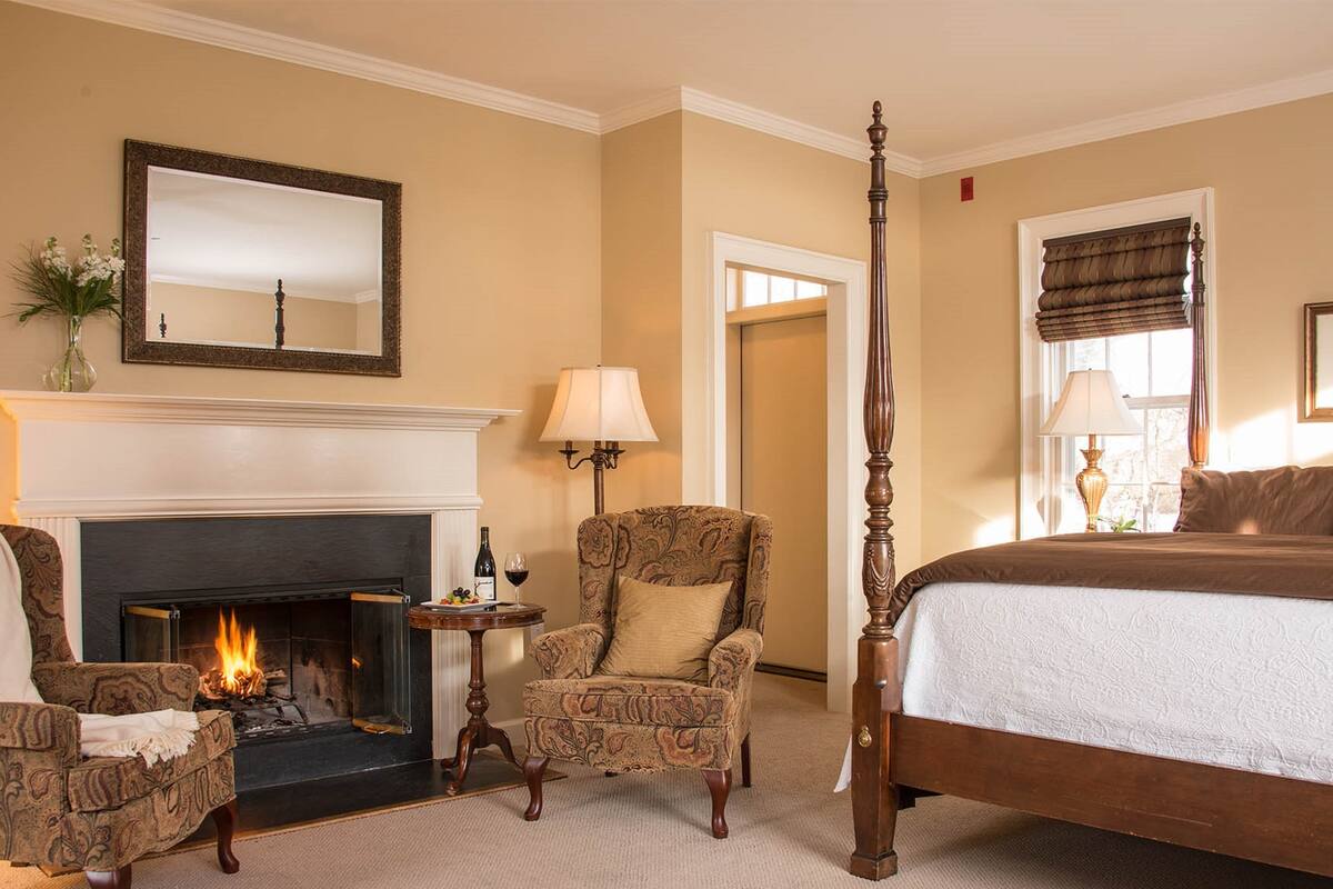 Quarto de hotel com cama de dossel, lareira, área de estar com uma poltrona e uma pequena mesa.