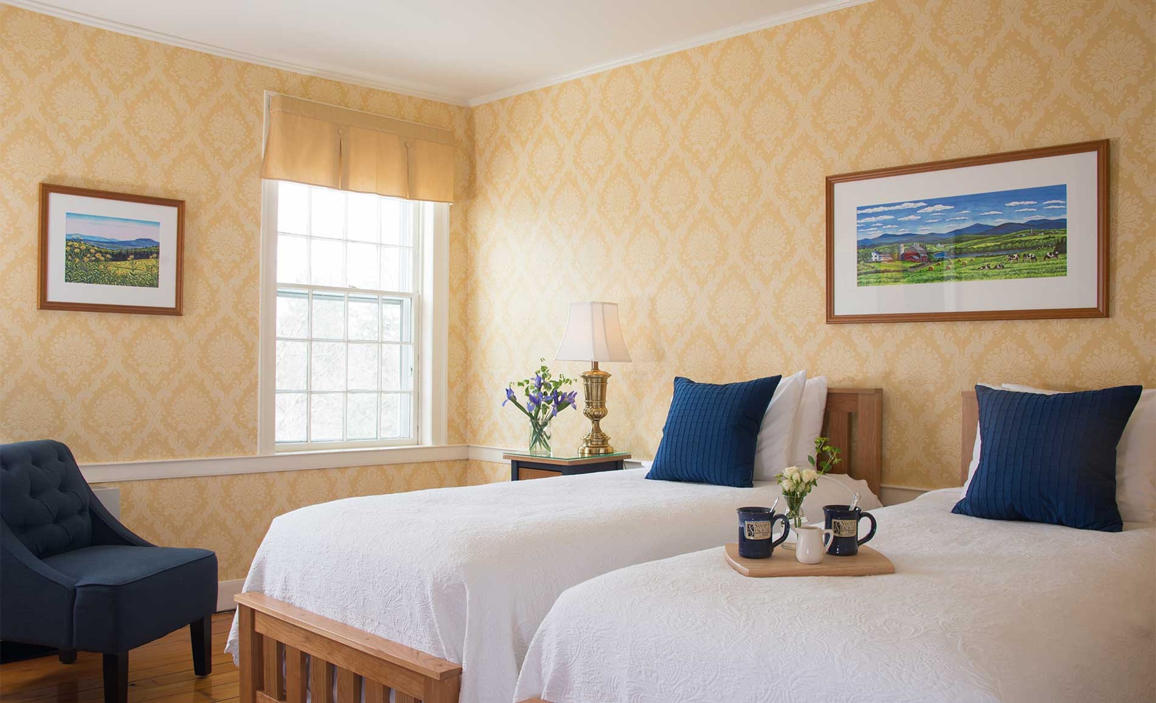 Habitación de hotel con dos camas, una ventana con cortinas, una lámpara y cuadros en las paredes.