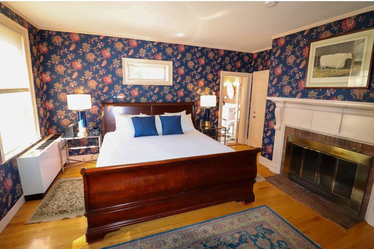 Quarto com cama grande, papel de parede floral, lareira e quadro na parede.
