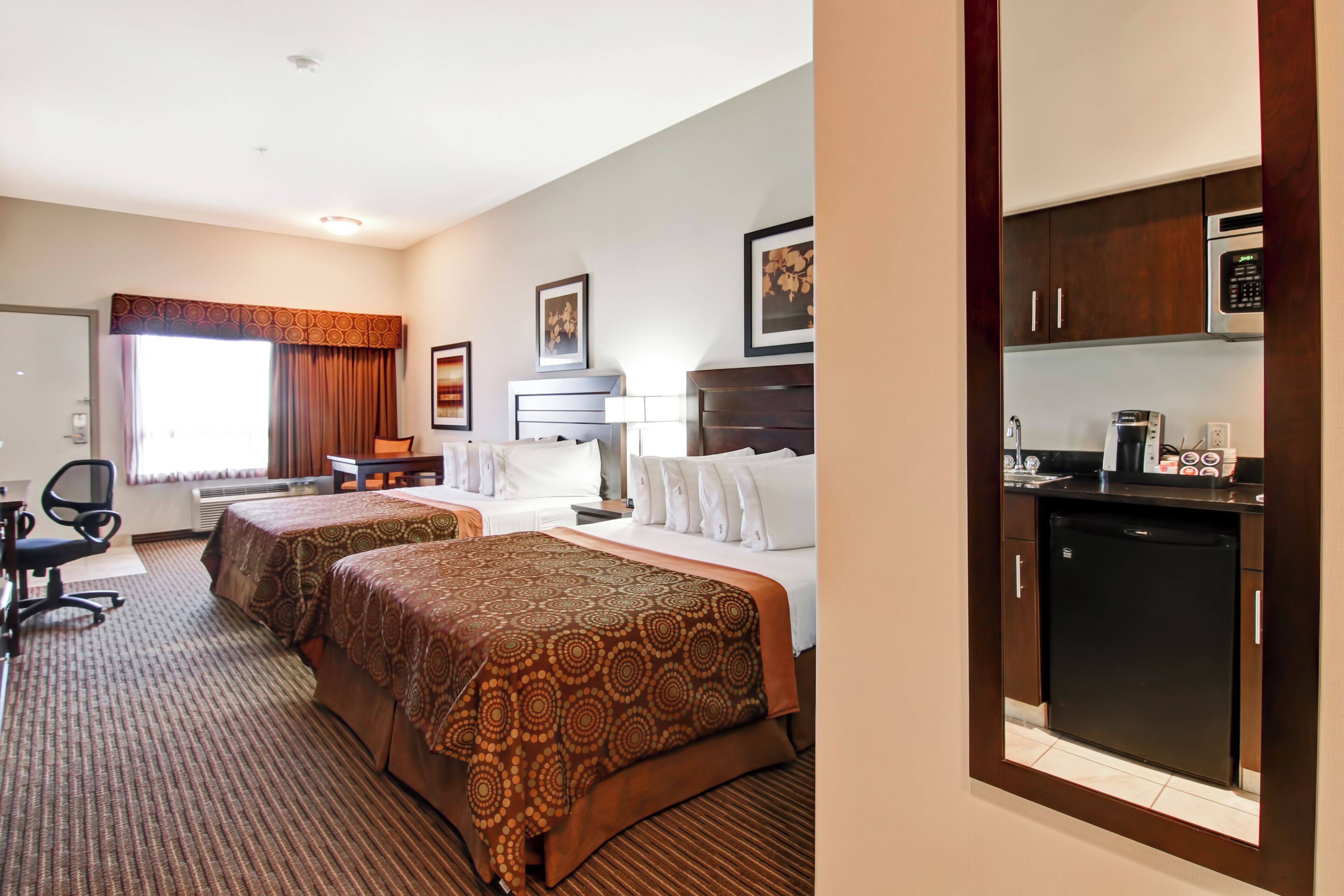 A hotel room with two beds, a desk, a chair, a desk lamp, a refrigerator, and a microwave.