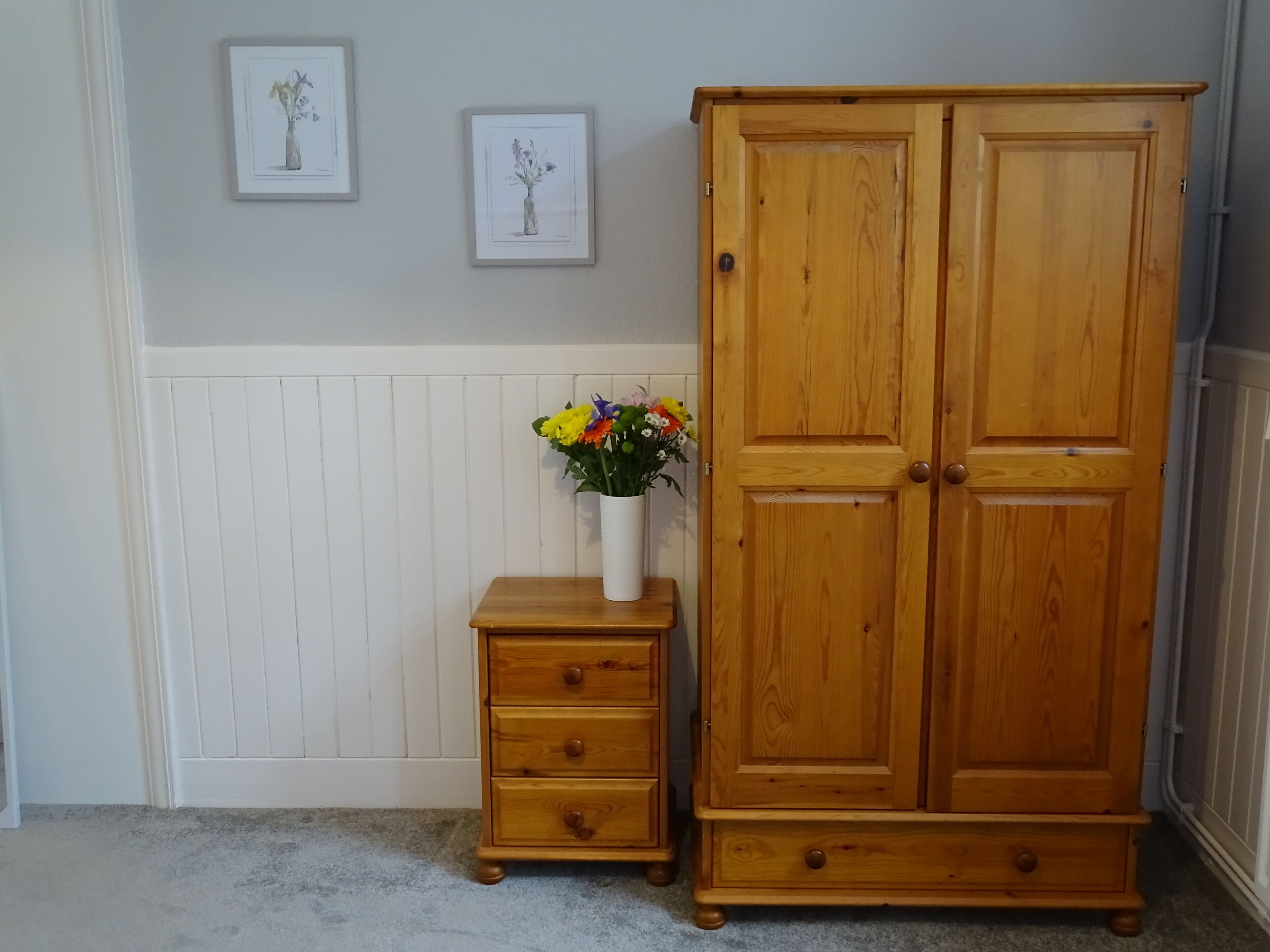 A wooden wardrobe with two doors, a small wooden nightstand with drawers, and a vase with flowers on the nightstand.