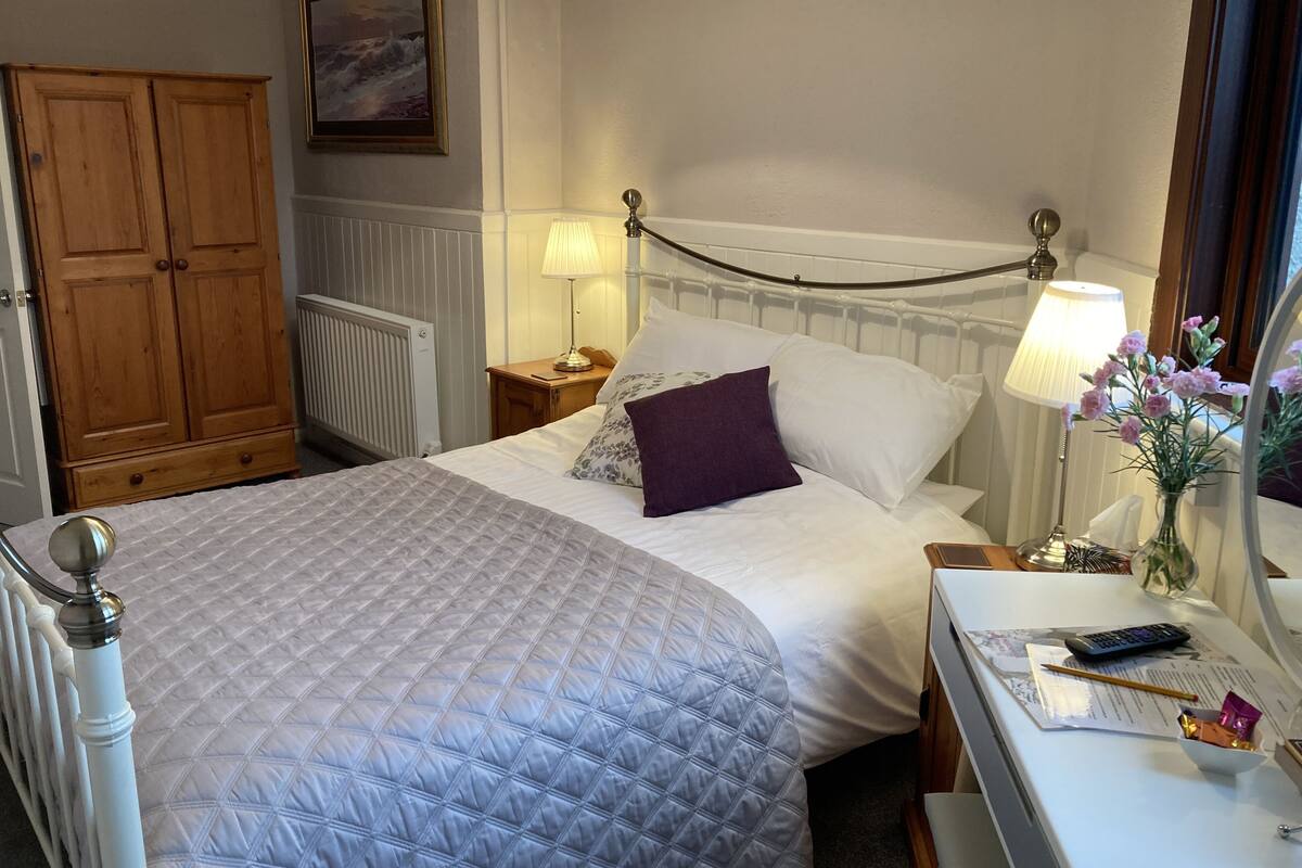 A neatly made bed with a quilted cover, a bedside lamp, a nightstand with a vase of flowers, and a wooden wardrobe.
