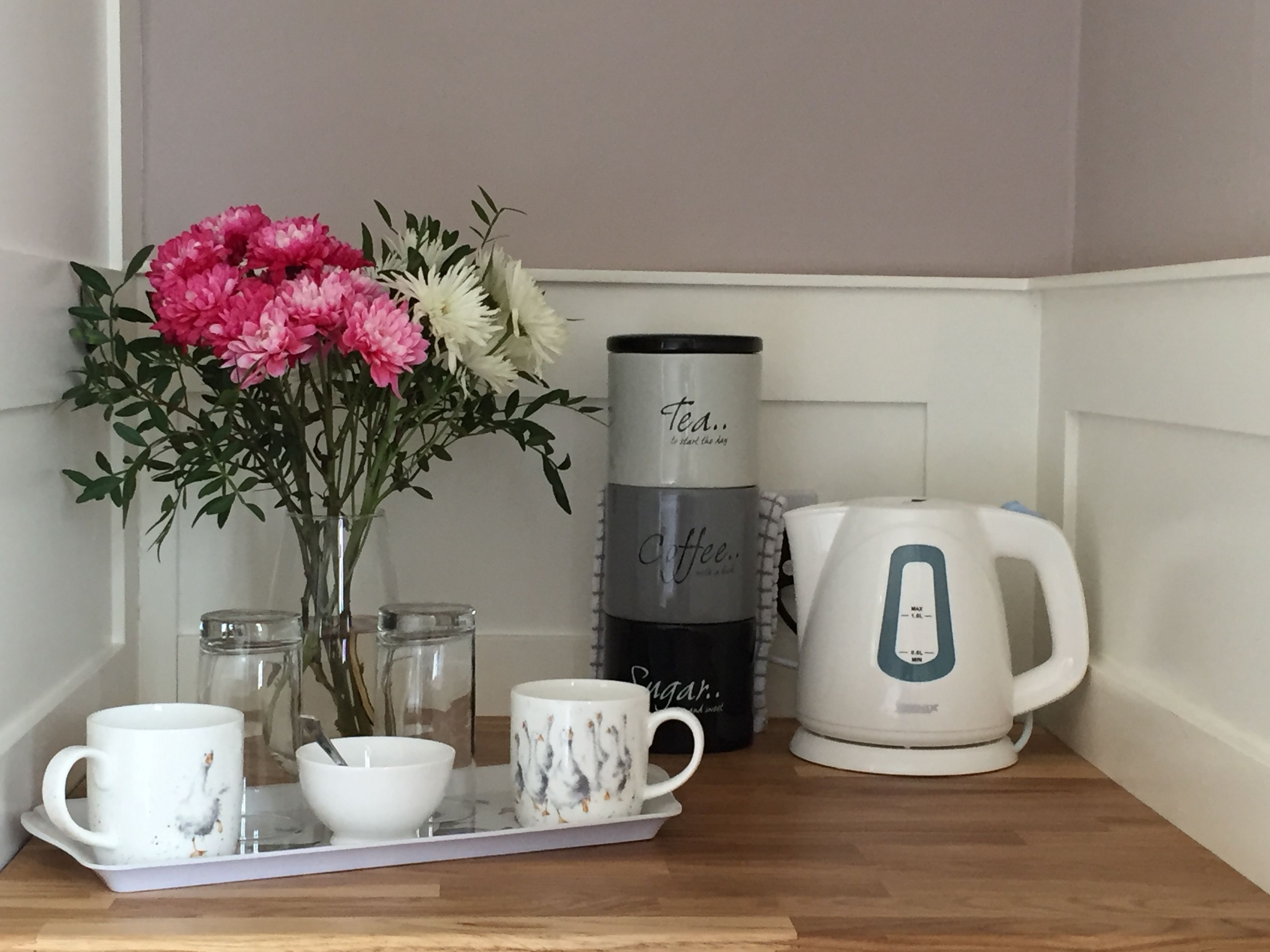 A kitchen counter with a tea kettle, a coffee maker, a mug, a bowl, and a tray with a vase of flowers.