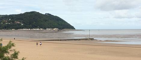 A beach with a sandy shore, a hill with greenery, and a cloudy sky.