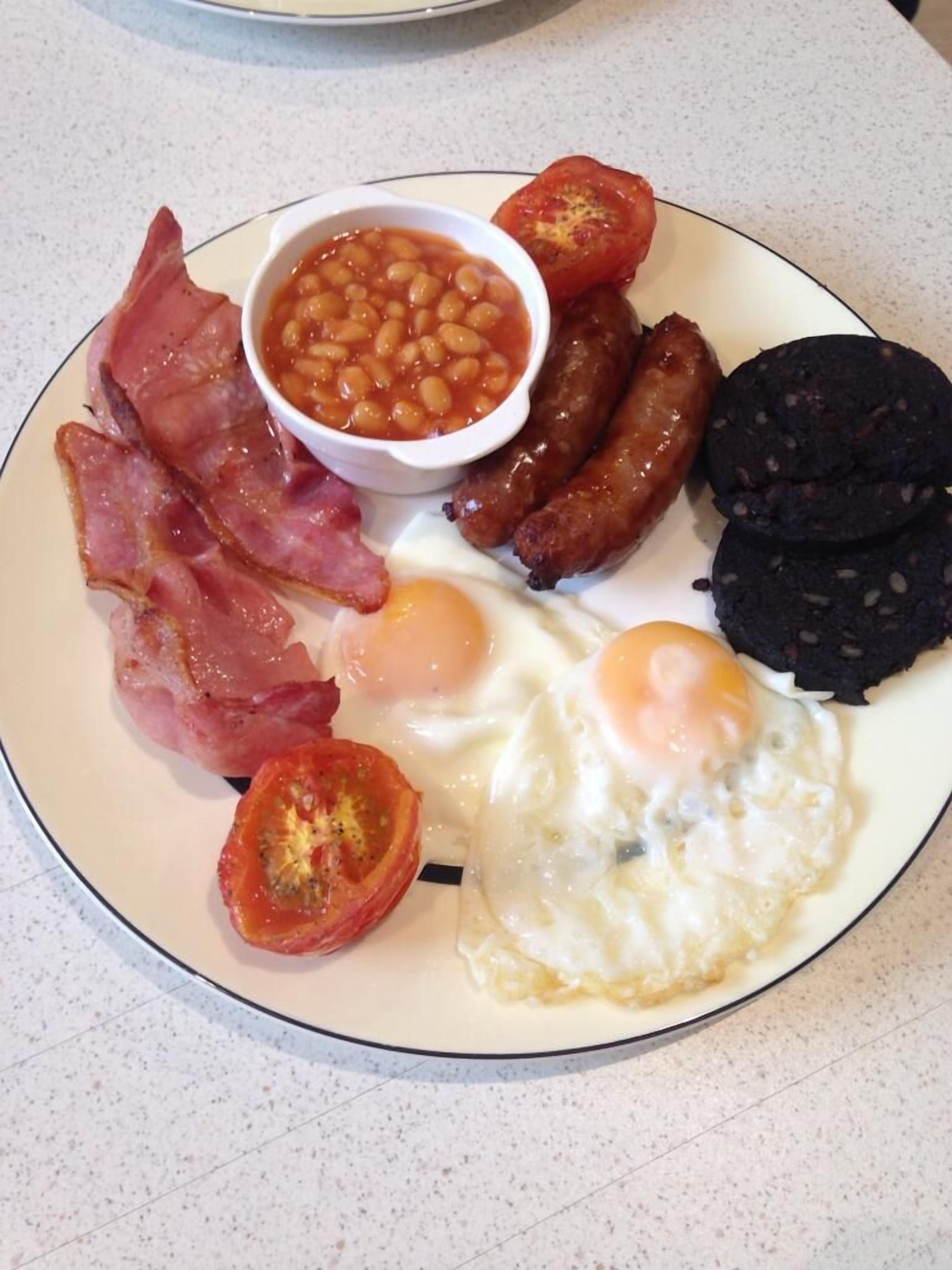 A plate of breakfast food including eggs, bacon, sausages, black pudding, baked beans, and tomatoes.