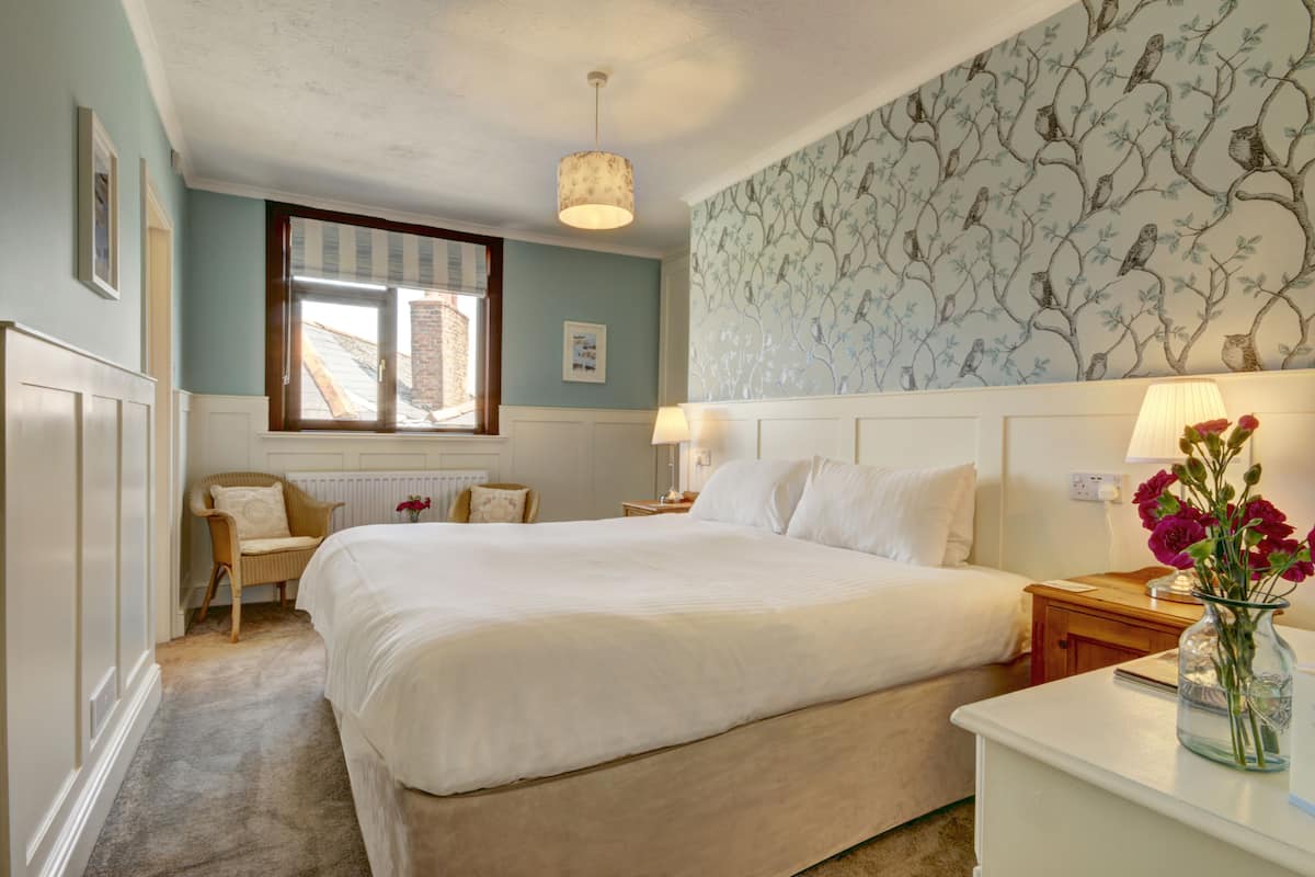 A bedroom with a large bed, a window, a chair, a nightstand, and a wall with a tree pattern.
