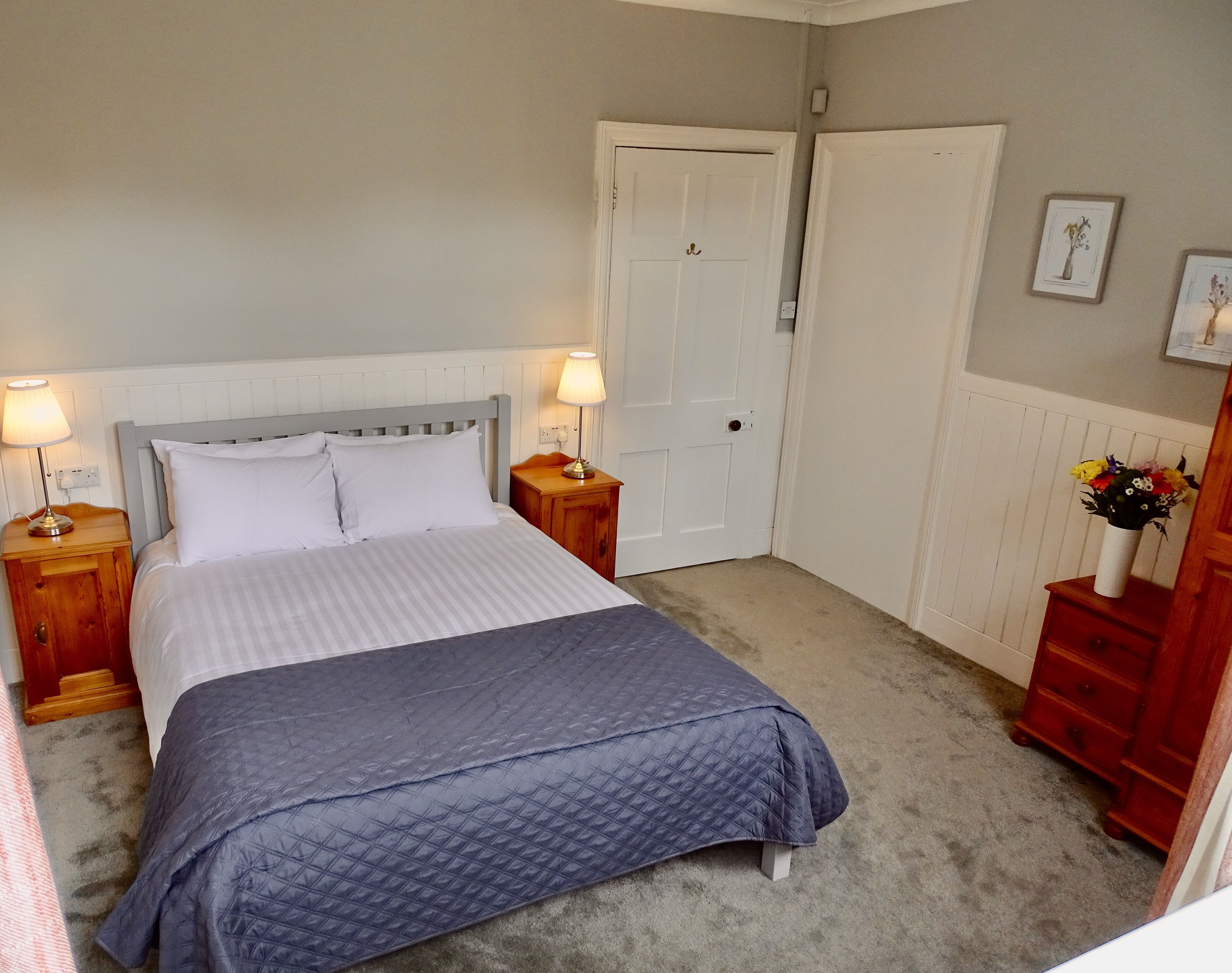 A bedroom with a bed, two bedside tables with lamps, a door, a dresser, and a wall with framed pictures.
