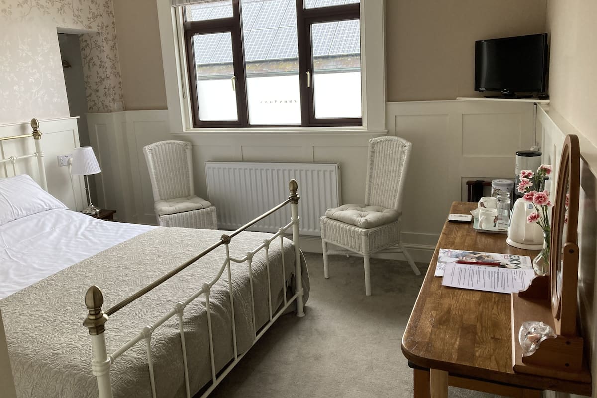 A hotel room with a bed, two chairs, a wooden table with a newspaper, a coffee maker, and a television on the wall.