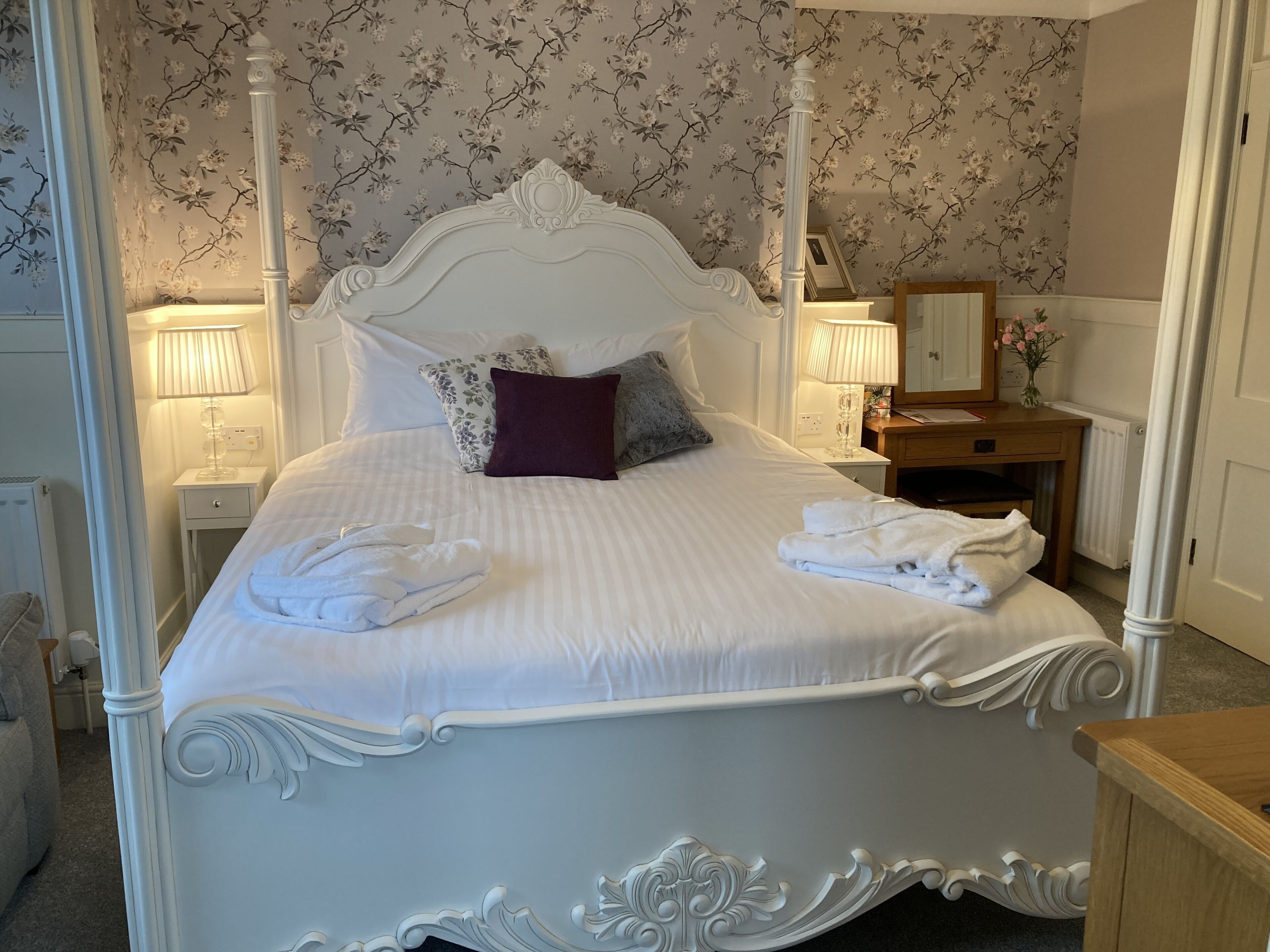 Una cama con dosel, con cabecera y piecera blancos, flanqueada por dos mesitas de noche con lámparas. Un aparador de madera se encuentra a la derecha de la cama.