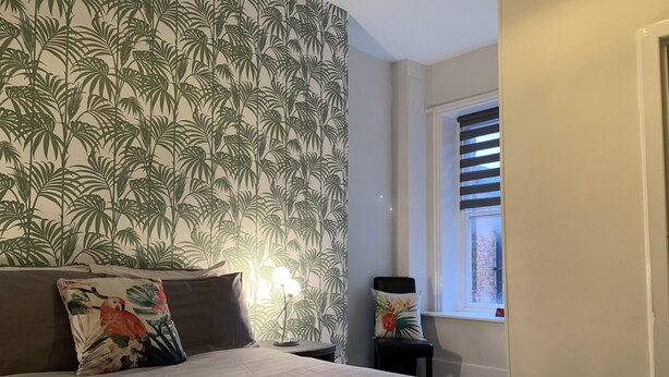 Ett hotellrum med en säng, två kuddar, ett nattduksbord, en lampa, ett fönster med persienner och utsikt över omgivningen.