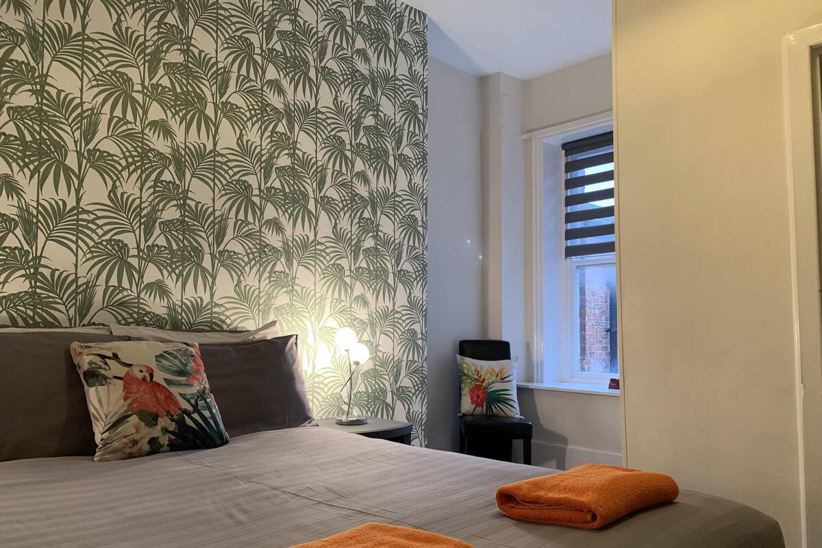 A hotel room with a bed, two pillows, a nightstand, a lamp, a window with blinds, and a view of the outside.