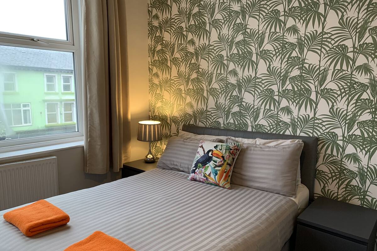 A neatly made bed with striped bedding, two orange towels, a wall with a tropical leaf pattern, and a window with curtains.