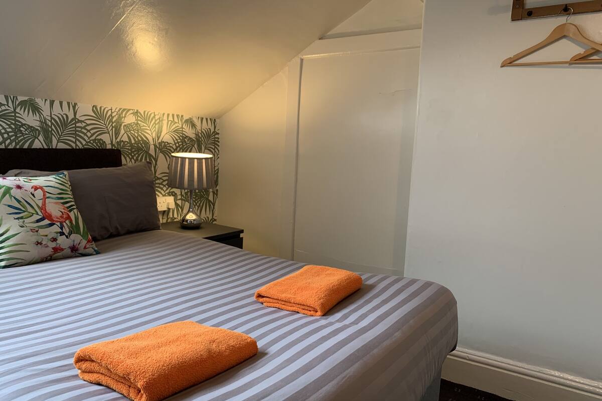 A neatly made bed with striped bedding and two folded orange towels. A lamp with a striped lampshade is on a bedside table, and a floral pillow is on the bed.