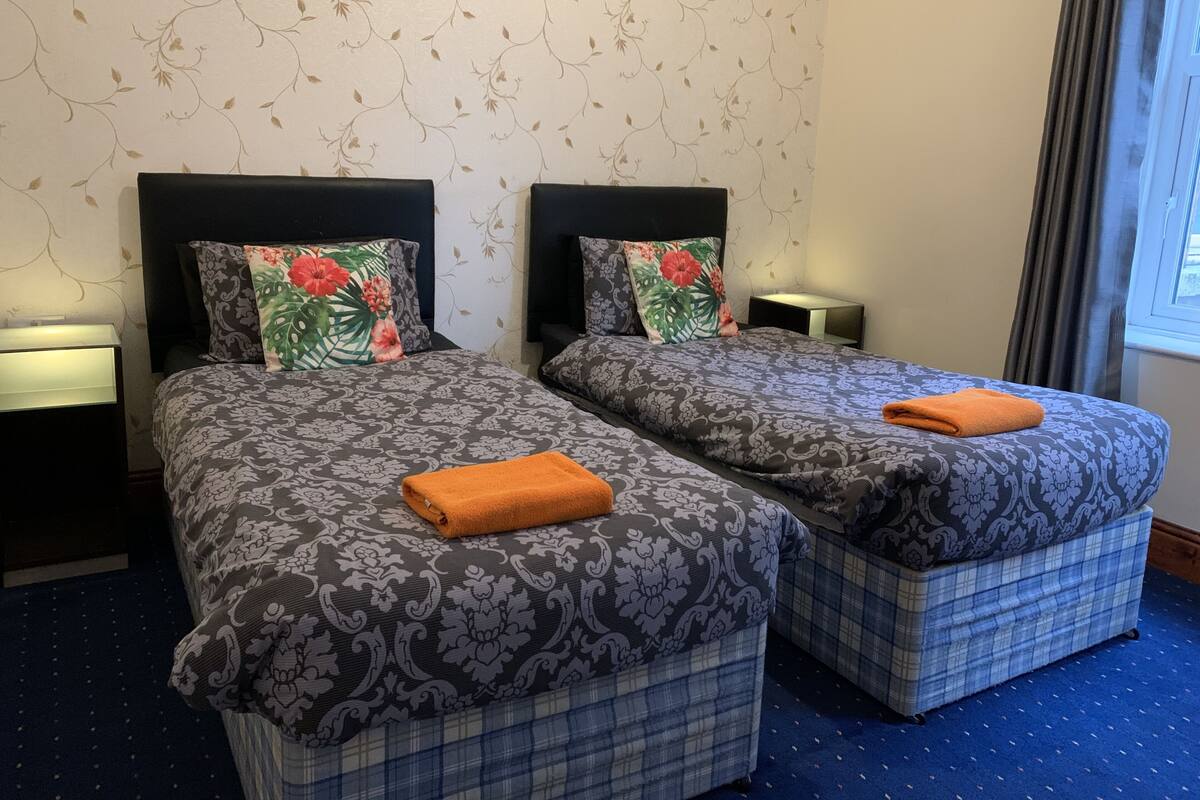 A hotel room with two beds, each with a patterned bedspread and matching pillows, a nightstand with a lamp, and a window with curtains.