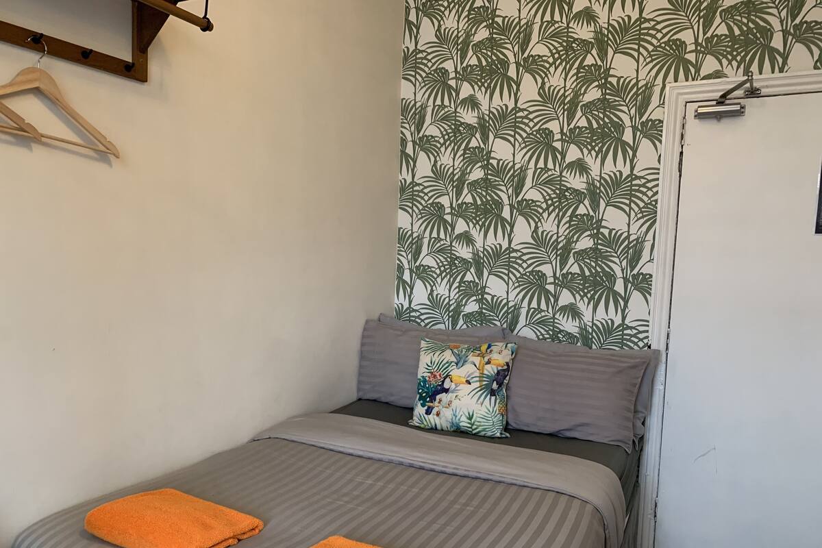A single bed with grey bedding and two orange towels, a wooden hanger, and a door with a plant-themed wallpaper.