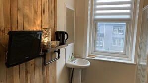 Ett badrum med en väggpanel av trä, en väggmonterad tv, en hylla med en vattenkokare och koppar, ett handfat och ett fönster med persienner.