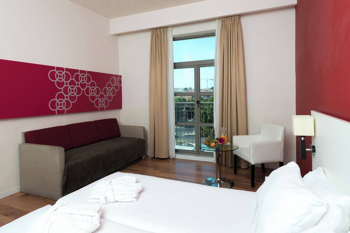 A hotel room with a bed, a sofa, a chair, a small table with a fruit bowl, and a window with curtains.