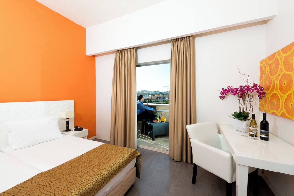 A hotel room with a bed, a desk, a chair, a table with wine bottles, a vase with flowers, and a view of a cityscape through the window.