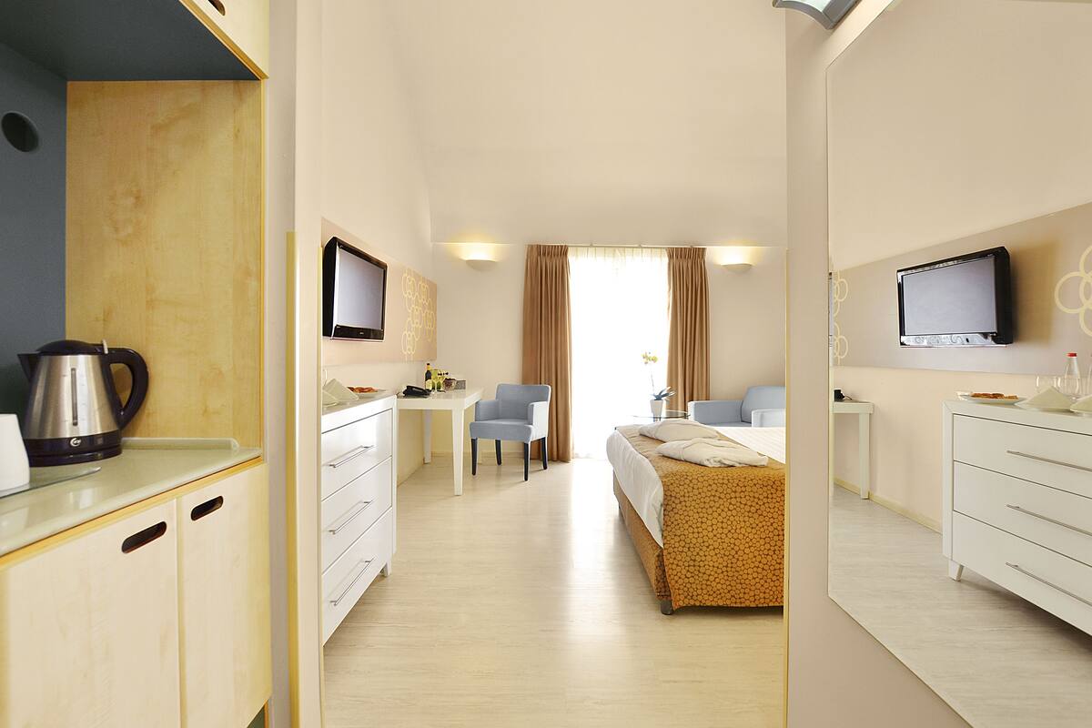 A hotel room with a bed, a desk, a chair, a TV, and a kitchenette.