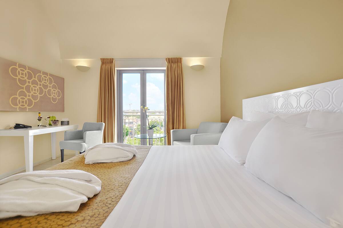 A hotel room with a bed, two armchairs, a desk, and a view of the city through large windows.