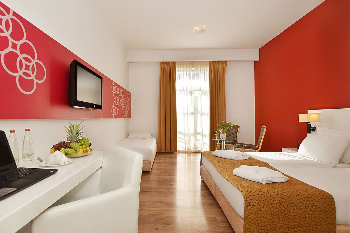 A hotel room with a desk, laptop, fruit bowl, two beds, a TV, and a window with curtains.
