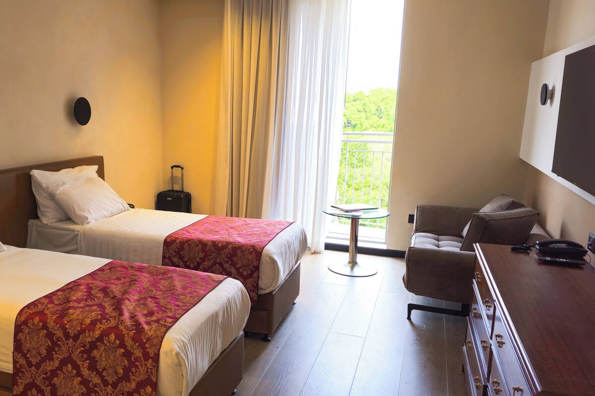 A hotel room with two beds, a desk, a chair, a small table, and a view of greenery outside.