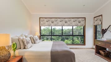 A bedroom with a large window, a bed with a beige and brown bedspread, a bedside table with a lamp, and a nightstand with a plant.
