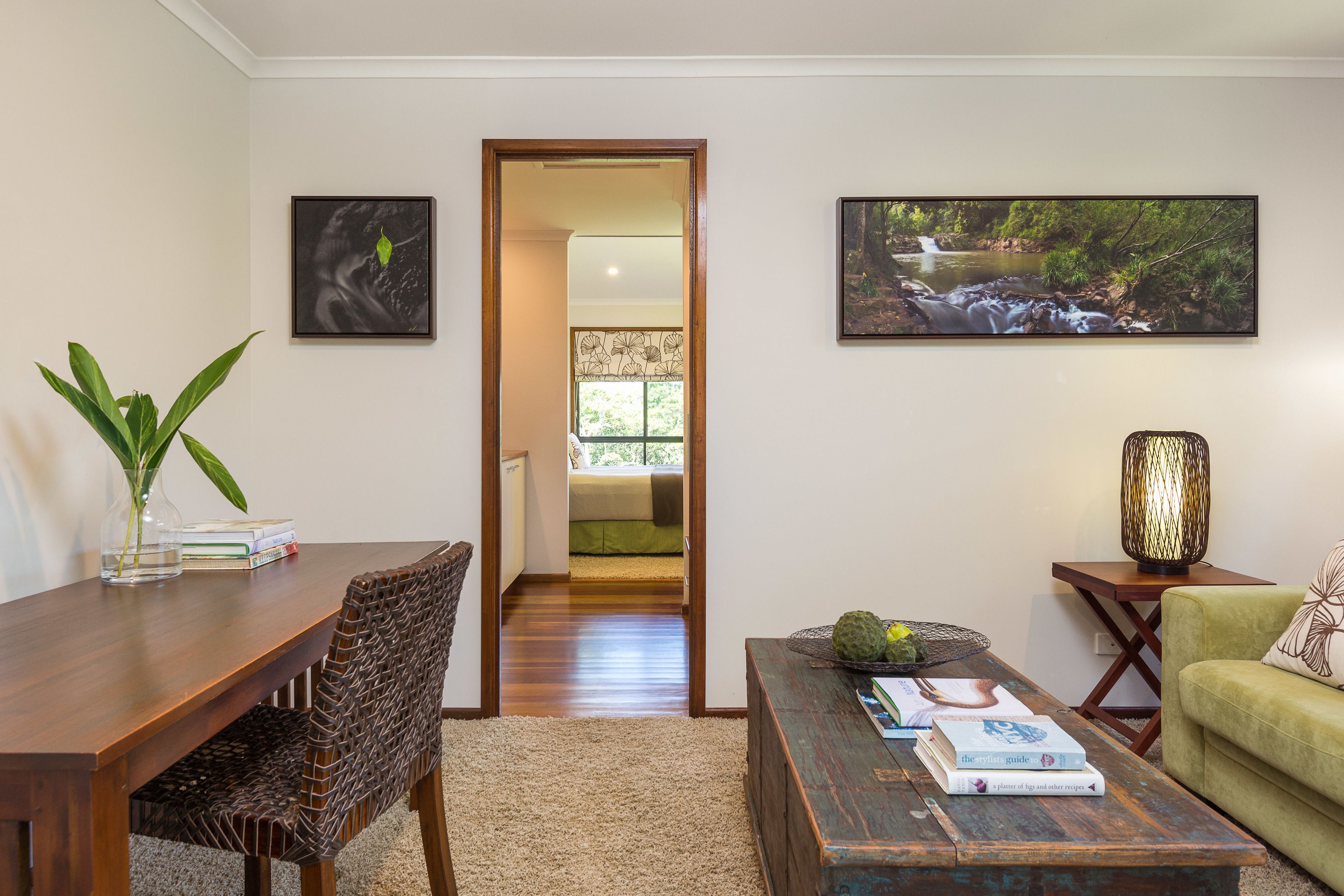 A living room with a wooden dining table, wicker chairs, a coffee table with books, a green sofa, and two framed pictures on the wall.
