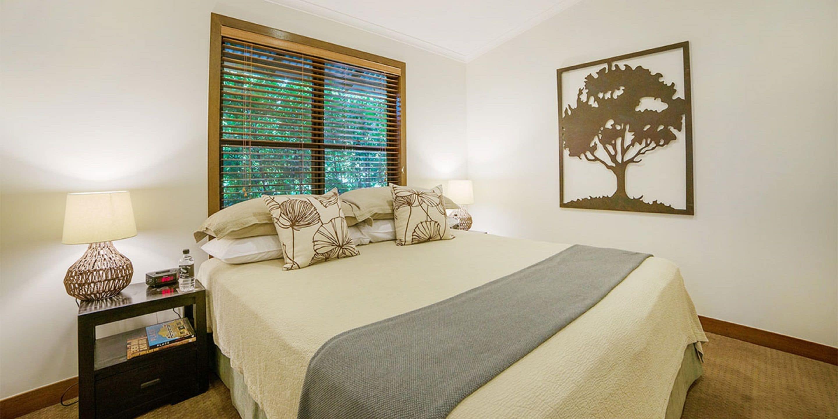 Cama bem arrumada com almofadas florais, abajur e quadro com uma árvore na parede.