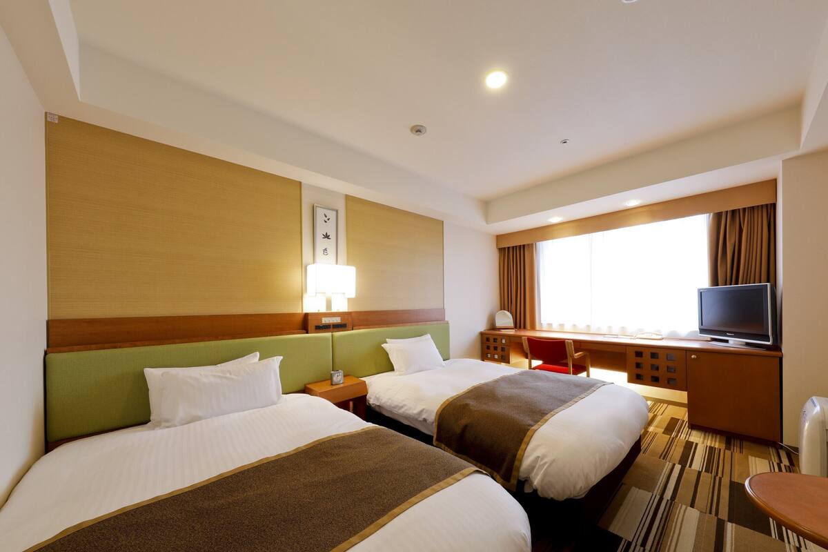 A hotel room with two beds, a desk, a chair, a TV, and a window with curtains.