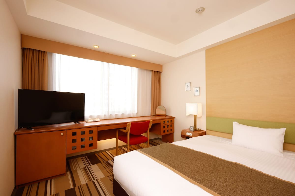 A hotel room with a bed, a desk with a television, a chair, a bedside table with a lamp, and a window with curtains.