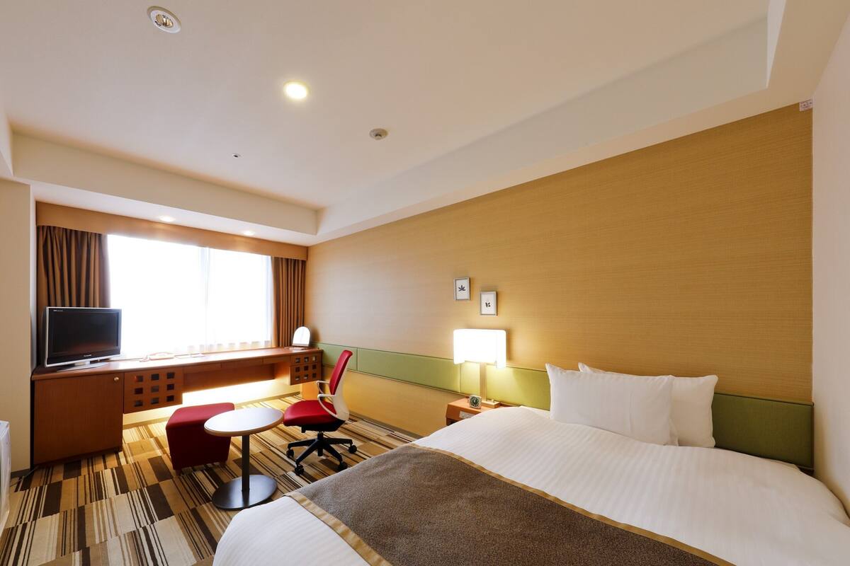 A hotel room with a bed, a desk with a television, a chair, a lamp, and a window with curtains.